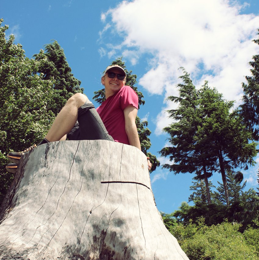 A picture of Peter sitting on a stump with sunglasses on and trees in the background